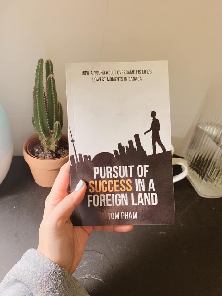 A hand holding the Pursuit of Success in a Foreign Land paperback beside a small cactus